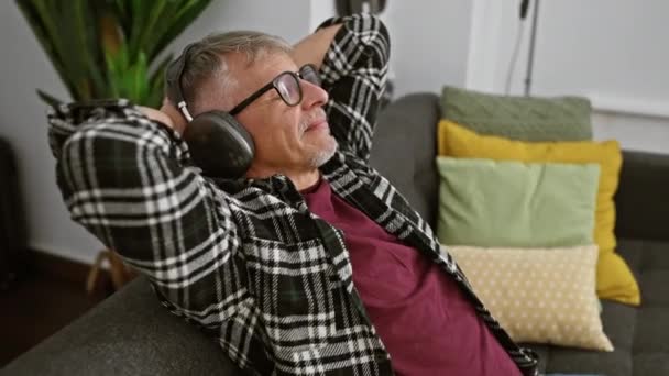 Расслабленный Пожилой Человек Наслаждается Музыкой Наушниках Сидя Дома Диване Окружении — стоковое видео