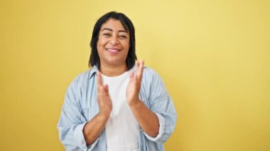 Gülümseyen olgun bir İspanyol kadın, sarı bir duvarın önünde el çırpıyor, günlük açık mavi kıyafetler giyiyor..