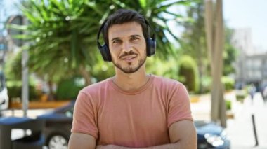 Kulaklık takan sakallı, gülümseyen genç adam şehir parkında güneşli bir günün tadını çıkarıyor..