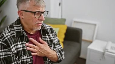 Gri saçlı bir adam oturma odasında otururken göğüs ağrısı çekiyor. Gözlük ve ekose gömlek takıyor..