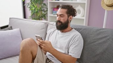 Modern oturma odasındaki koltukta akıllı telefon kullanan yakışıklı sakallı adam.