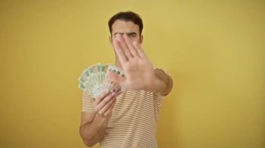 Kendine güveni tam genç İspanyol bir adam sıkı bir duruş hareketi yapıyor, elinde para tutuyor - limitlere karşı ciddi bir savunma ve izole edilmiş sarı arka plana yasaklı yatırımlar