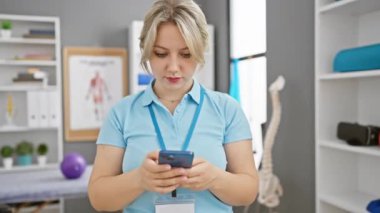 Üniformalı sarışın sağlık uzmanı modern rehabilitasyon kliniğinde akıllı telefon kullanıyor.