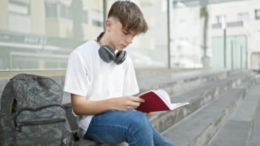 Genç bir adam, şehir ortamında öğrenci hayatını tasvir eden kulaklıklar ve sırt çantasıyla merdivenlerde otururken kırmızı bir kitap okuyor..