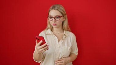 Şaşırmış genç sarışın kadın, ağzı açık, akıllı telefondaki şok edici mesaj karşısında dehşete düşmüş, korkmuş, güvensiz bir yüz ifadesi izole edilmiş kırmızı arka planda