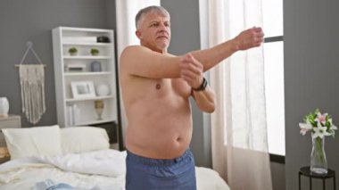 Orta yaşlı bir adam kollarını modern bir yatak odasında uzatıyor, sağlıklı bir sabah rutinini tasvir ediyor.