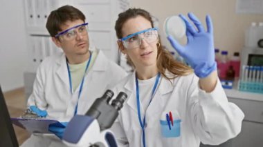 Bir kadın bilim adamı, erkek arkadaşı laboratuvar ortamında notlar alırken bir örneği inceliyor.