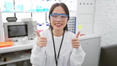 Neşeli genç Çinli bilim kadını, laboratuvarda başarıya ve pozitifliğe parmak basıyor! Zaferinin tadını çıkarırken mutluluk saçıyor..