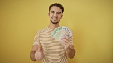 Neşeli, genç İspanyol adam büyük gülümsüyor, başparmağıyla onay işareti yapıyor. Tamamen yalıtılmış sarı arka planda, bu adam pozitif, başarılı yaşam enerjileri yayıyor.