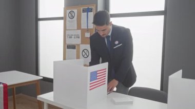 Yakışıklı genç bir adam oy pusulası ve bayrağıyla bir Amerikan seçim merkezinde oy veriyor.
