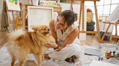 Genç İspanyol kadın sanatçı yerde oturuyor, sevgiyle gülümsüyor ve renkli bir sanat stüdyosunda sevimli köpeğini öpüyor.