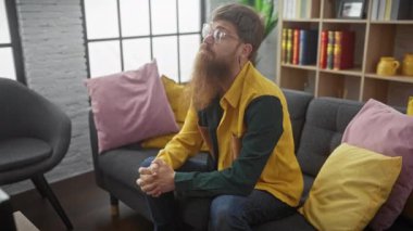 Gözlüklü sakallı bir adam, modern oturma odasındaki sarı ve pembe yastıklı gri bir koltukta düşünceli bir şekilde oturur..