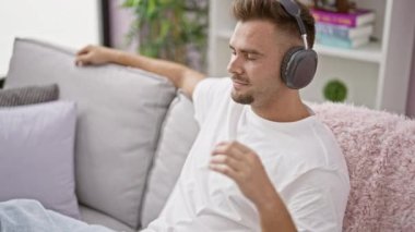 Neşeli genç bir adam, rahat bir oturma odasındaki kanepede dinlenirken kulaklıkla müzik dinlemekten hoşlanır..