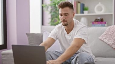 Odaklanmış İspanyol bir adam dizüstü bilgisayarda çalışıyor ve rahat bir oturma odasında telefonla konuşuyor.