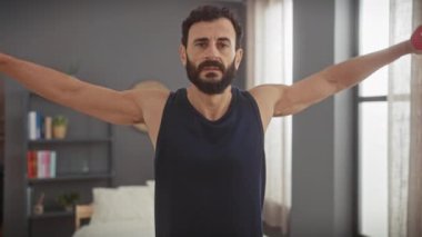 Orta yaşlı, sakallı, kolsuz bluz giyen, temiz bir yatak odasında dambıllarla egzersiz yapan bir adam.