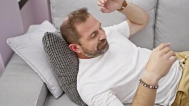 Rahat, orta yaşlı, sakallı bir adam rahat bir evde, rahat ve huzurlu bir koltukta uzanıyor..