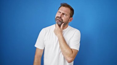 Beyaz tişörtlü, beyaz saçlı, orta yaşlı bir adam. Düşünce ifadeleri sergiliyor ve mavi arka plana karşı bir düşüncesi var..