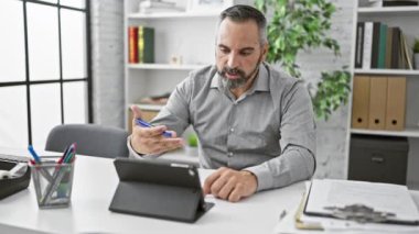 Gri saçlı ve sakallı olgun bir adam modern bir ofiste tabletle çalışmaya odaklanmış.