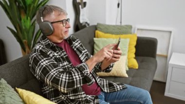 Gri saçlı adam, rahat bir oturma odasında akıllı telefon kullanırken kulaklıkla müzik dinlemekten hoşlanıyor..