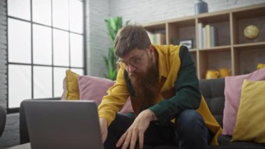 Gözlüklü ve sarı ceketli sakallı adam modern bir oturma odasında dizüstü bilgisayar kullanıyor.