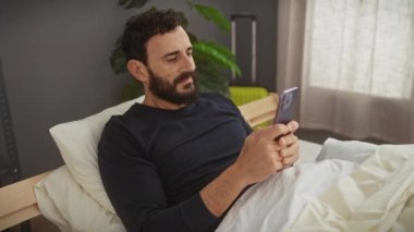 Sakallı adam rahat bir yatak odası ortamında dinlenirken akıllı telefon kullanıyor. Huzuru ve gündelik yaşamı ifade ediyor..