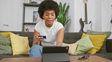 Gülümseyen Afrikalı Amerikalı kadın rahat oturma odasında online alışveriş için kredi kartı kullanıyor..