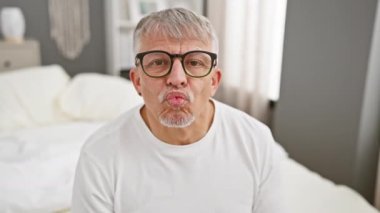 Neşeli orta yaşlı, gri saçlı bir adam kameraya aşk dolu bir öpücük yolluyor, pijamalarıyla çok tatlı görünüyor. Yatak odasının rahatlığından seksi pozitifliğini gösteriyor..