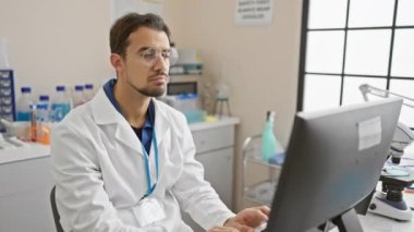 Beyaz laboratuvar önlüklü, modern bir laboratuvarda bilimsel ekipmanlarla bilgisayar başında çalışan ciddi bir İspanyol..