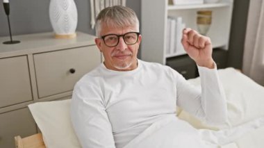 Orta yaşlı, gri saçlı, yatak odasında pijama giyen, parmaklarıyla küçük beden işareti yapan kendinden emin bir adam. Küçük ölçü, büyük kavram!