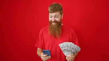 Neşeli genç kızıl saçlı adam kazandıklarının üzerine ışıldıyor, dolarları ve akıllı telefonu izole edilmiş kırmızı arka planda tutuyor.!