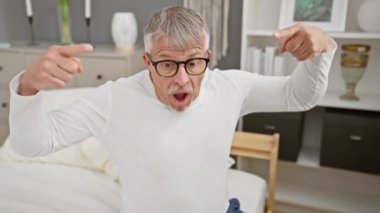 Şaşırmış orta yaşlı, gri saçlı, pijama giyen, neşe içinde aşağıya bakan, yatak odasının rahatlığında inanılmaz bir reklam veren bir adam.