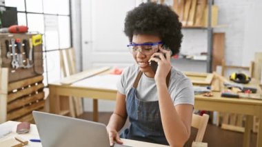 Marangoz stüdyosunda çalışan, güvenlik gözlüğü takan, telefon ve dizüstü bilgisayar kullanan Afrikalı Amerikalı bir kadın..