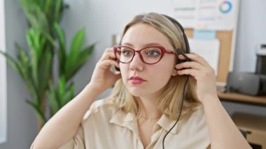 Odaklanmış, sarı saçlı, gözlüklü ve modern bir ofis ortamında kulaklık takan genç bir kadın..