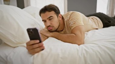Sakallı yakışıklı, genç, İspanyol bir adam akıllı telefonunu kullanarak yatağında uzanıyor ve modern ev hayatının bir anını yakalıyor..