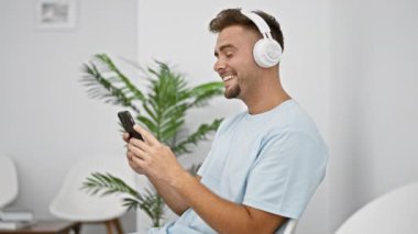 Gülümseyen İspanyol bir adam kulaklık takıyor ve modern beyaz bir odada sandalye ve bitkilerle akıllı telefon kullanıyor.