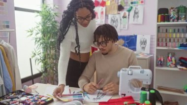 Bir kadın ve bir erkek dikiş makinesi, kumaş, eskiz ve iplik arasında bir terzi atölyesinde işbirliği yapıyor.