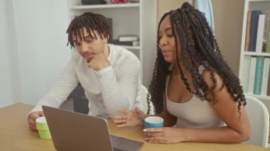 Gülümseyen bir çift evde bir video görüşmesi yapıyor, dizüstü bilgisayar, kahve fincanları ve arka planda bir kitaplık ile..