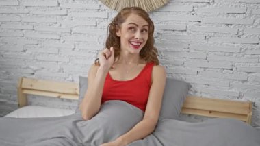 Heyecanlı genç bir kadın, güvenli bir şekilde yatak odasında oturuyor, neşeli bir şekilde bir parmağıyla zekice bir fikri işaret ediyor. Pijama giyiyor, güzel yüz ışıklarını takıyor ve gülümsüyor..
