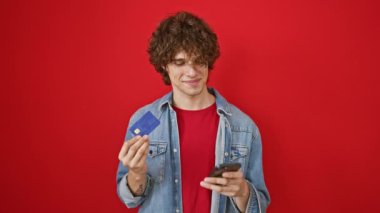 Kıvırcık saçlı gülümseyen genç adam kredi kartı ve akıllı telefonu kırmızı arka planda tutuyor ve modern ödeme yöntemlerini tasvir ediyor..