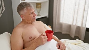 Olgun bir adam yatakta kahve içiyor, rahatlamış bir sabah evde rahatlık ve günlük rutini vurguluyor..