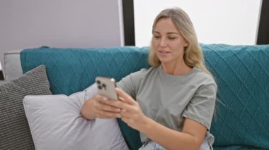 Sarışın kadın akıllı telefon kullanıyor ve turkuaz koltukta dinleniyor.