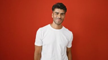 Beyaz tişörtlü, sakallı yakışıklı İspanyol bir adam, izole edilmiş kırmızı bir arka plana selam veriyor.