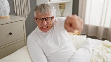 Öfkeli, kır saçlı, orta yaşlı bir adam, yatak odasında kızgın görünüyor, pijama giyiyor, seni işaret ediyor, kamera karşısında hoşnutsuz.