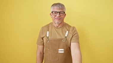 Gri saçlı, sarı izole edilmiş arka planda duran neşeli orta yaşlı adam. Gözlük ve önlük takıyor, yardım etmek, kabul etmek ve yaşlılara destek olmak için elini uzatıyor..