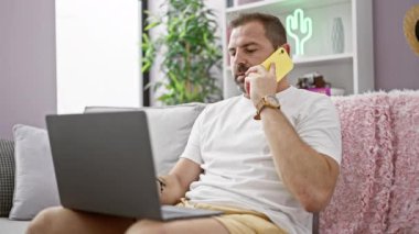Orta yaşlı, gri saçlı, modern bir oturma odasında akıllı telefondan konuşan bir adam.