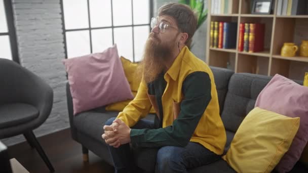 Homem Barbudo Óculos Senta Pensativamente Sofá Cinza Com Travesseiros Amarelos — Vídeo de Stock