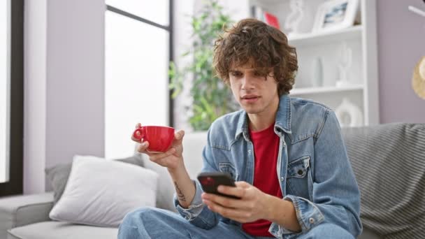 穿着粗斜纹棉布夹克的卷发年轻人 用智能手机 在现代客厅里拿着红色的杯子 — 图库视频影像