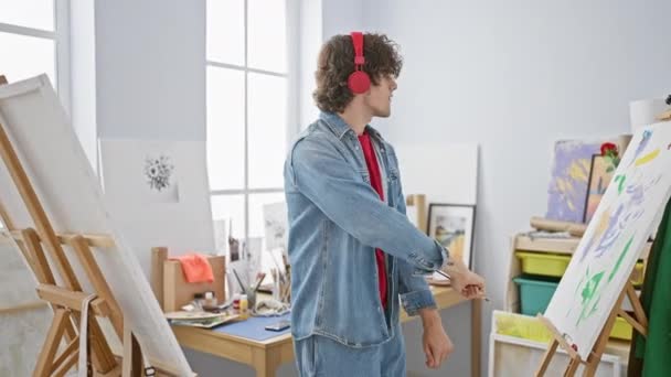 明るいアートスタジオのイーゼルにヘッドフォンを着用した若い大人男性 創造性とフォーカスを体現 — ストック動画