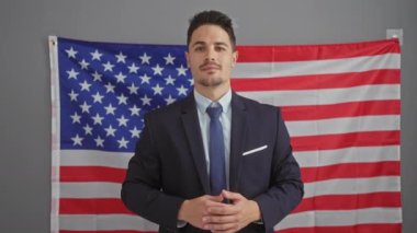 Takım elbiseli kendine güvenen İspanyol işadamı, bir ofis ortamında Amerikan bayrağı önünde kamerayı işaret ederek poz veriyor..