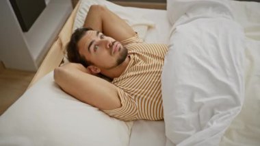 Düşünceli, genç, sakallı bir adam yatakta uzanıyor, parlak bir yatak odası ortamında düşünceli görünüyor..
